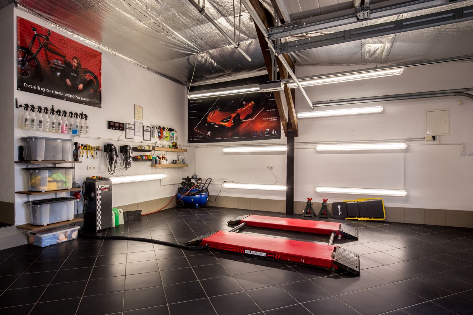 Stanowisko warsztatowe MTM Auto Detailing — platformy najazdowe do wyciszania, ściana z chemią detailingową, plakat brandowy, oświetlenie LED